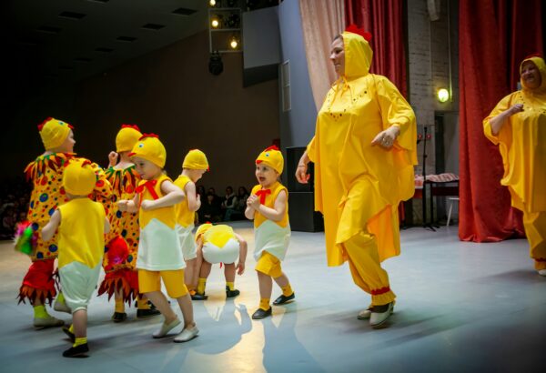 School children with two teachers all dressed as chickens performing on stage.