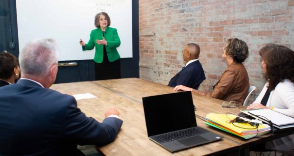 Teresa Zumwald in front of a white board teaching workshop attendees sitting at a table in a conference room.