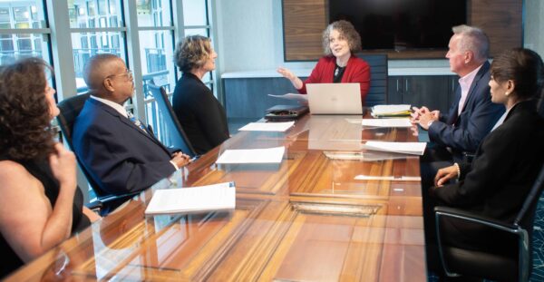 Teresa Zumwald speaking at a conference table, addressing several business people seated around the table in a professional setting.