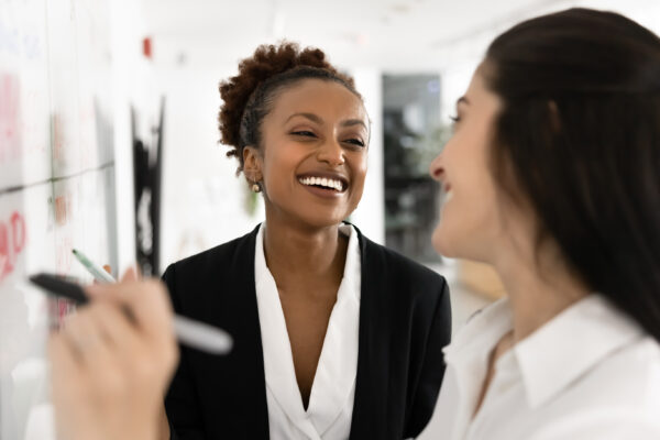 Professional woman smiling and conversing with a coworker who is writing on a whiteboard