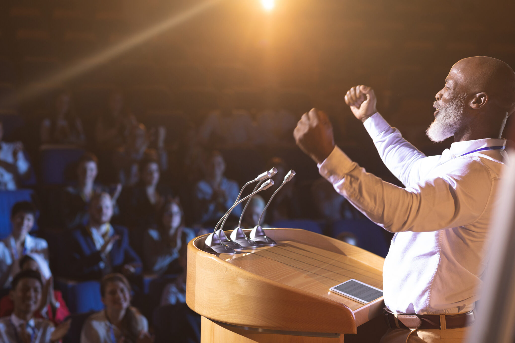 Side view of old African-American businessman standing near podium and giving speech to the audience in the auditorium